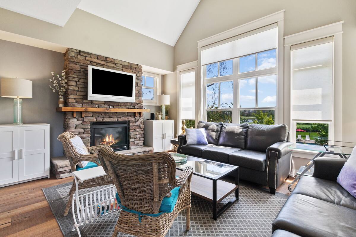 A spacious living room with a stone fireplace, comfortable seating, large windows, and a flat-screen TV.