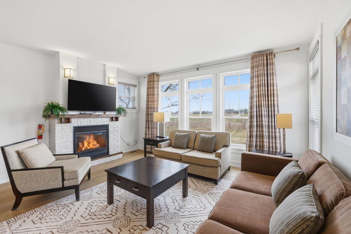 A living room with a fireplace, sofa, armchair, TV, and large windows.