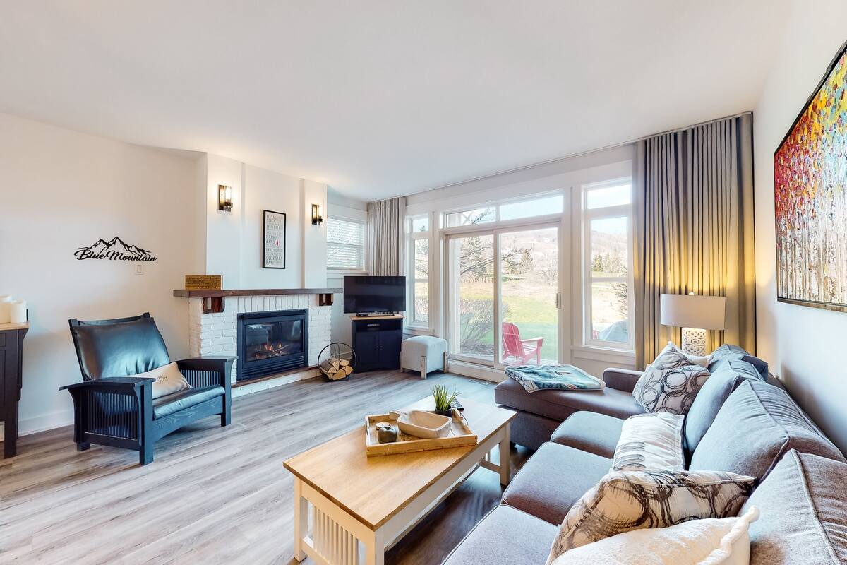 A modern living room with a fireplace, leather chairs, a wooden coffee table, and a large window with curtains.