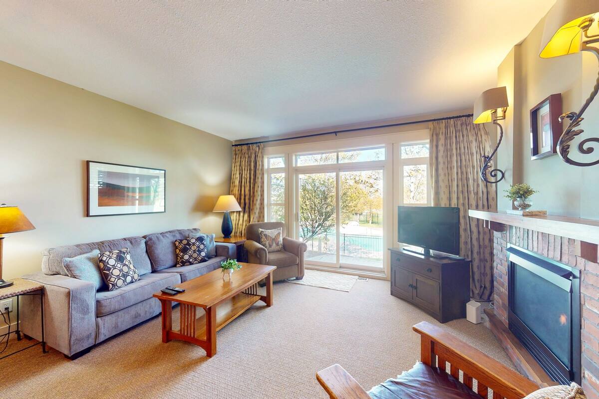 A living room with a sofa, armchair, coffee table, TV, fireplace, and large windows.
