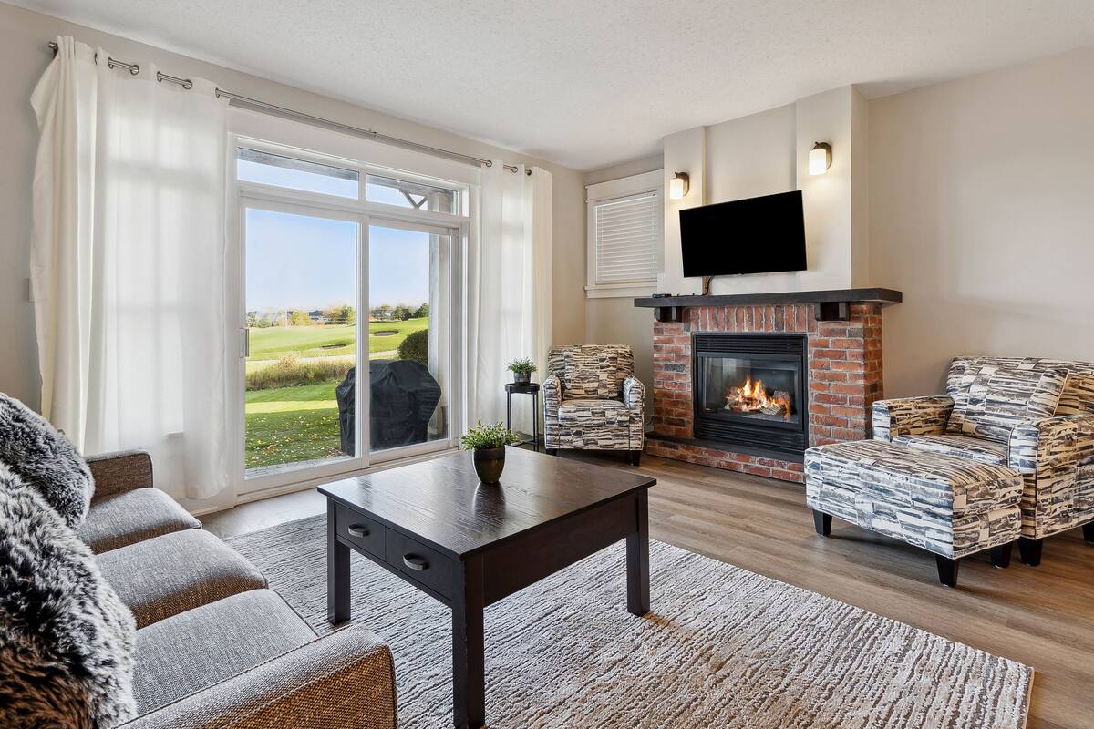 A living room with a fireplace, a sofa, a coffee table, and a view of a green landscape through large windows.