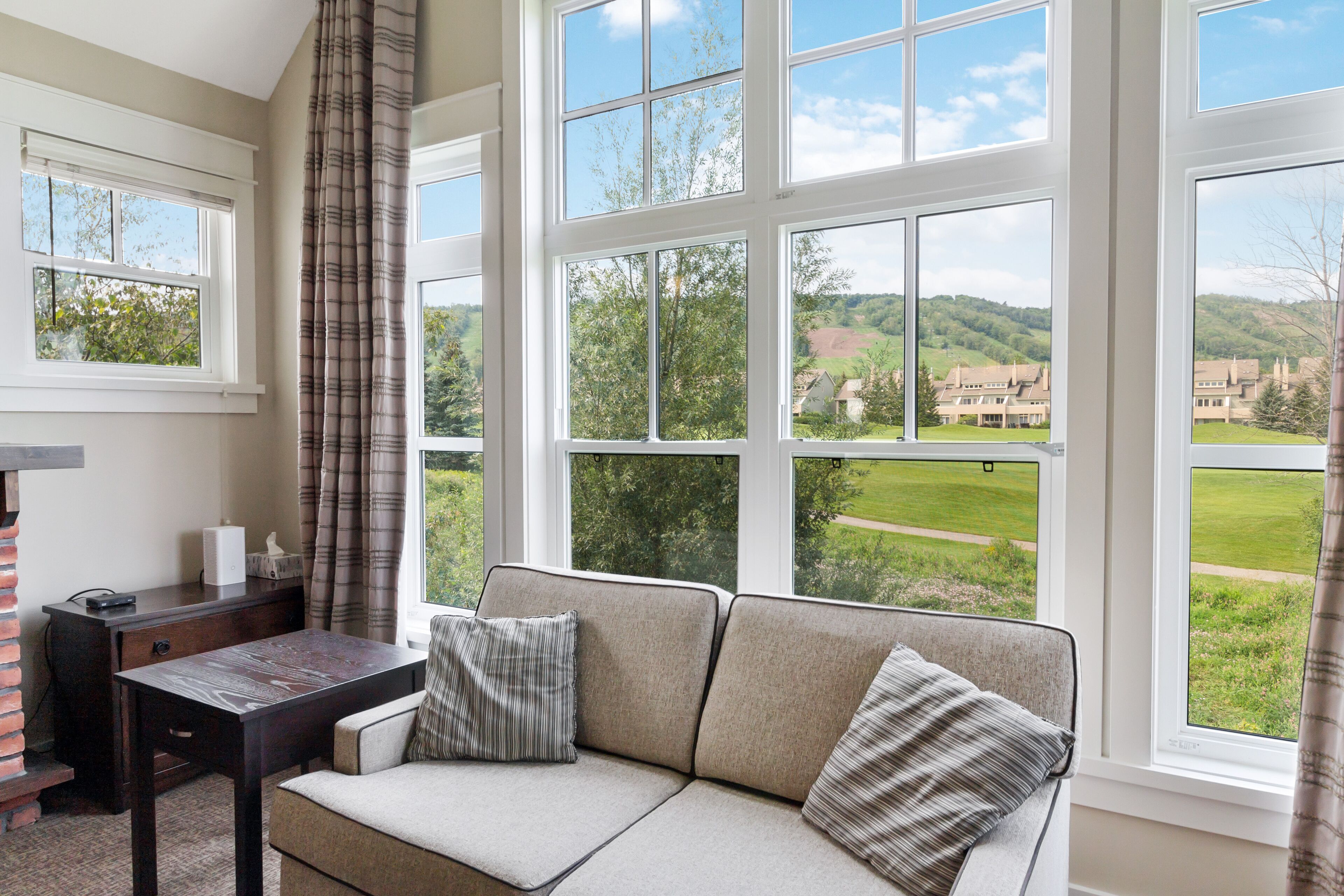 Habitación con sofá frente a grandes ventanales, un escritorio con lámpara y vistas a un paisaje verde.