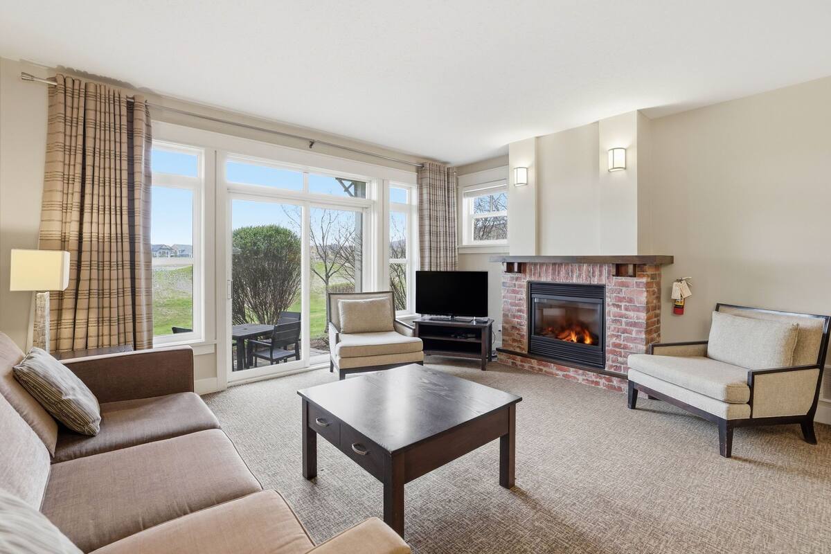 A living room with a sofa, armchair, coffee table, TV, fireplace, and large windows.