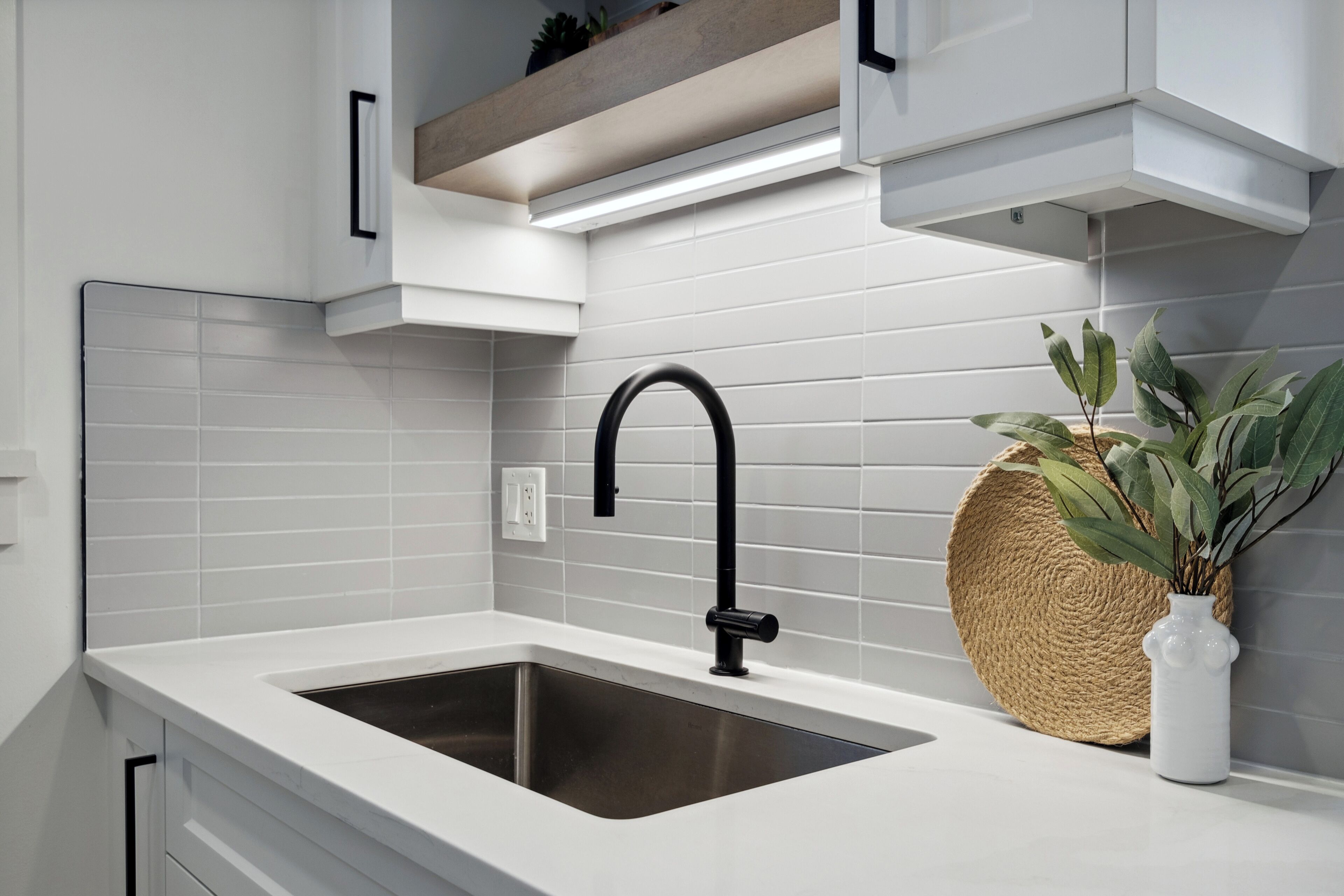 A modern kitchen with a white countertop, a stainless steel sink, and a black faucet. There is a woven basket and a white vase with a plant on the counter.