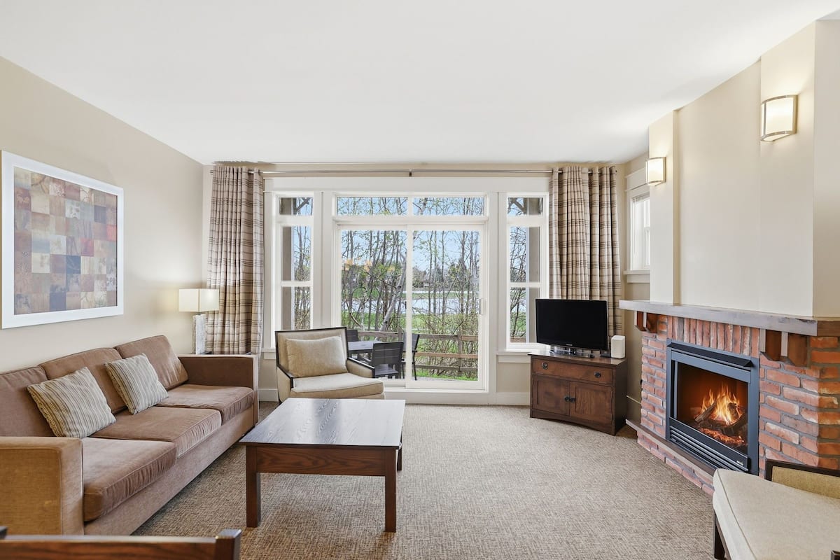 A living room with a sofa, coffee table, television, fireplace, and large windows.