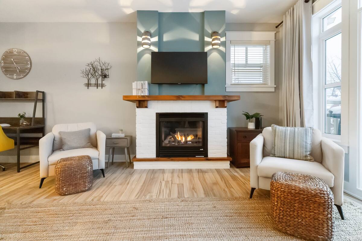 A living room with a fireplace, two armchairs, a coffee table, a TV mounted above the fireplace, and a clock on the wall.