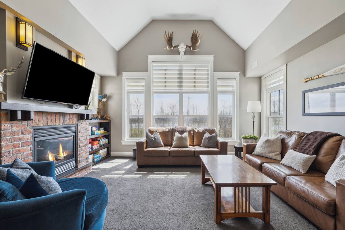 A cozy living room with a fireplace, leather sofas, a flat-screen TV, and large windows.