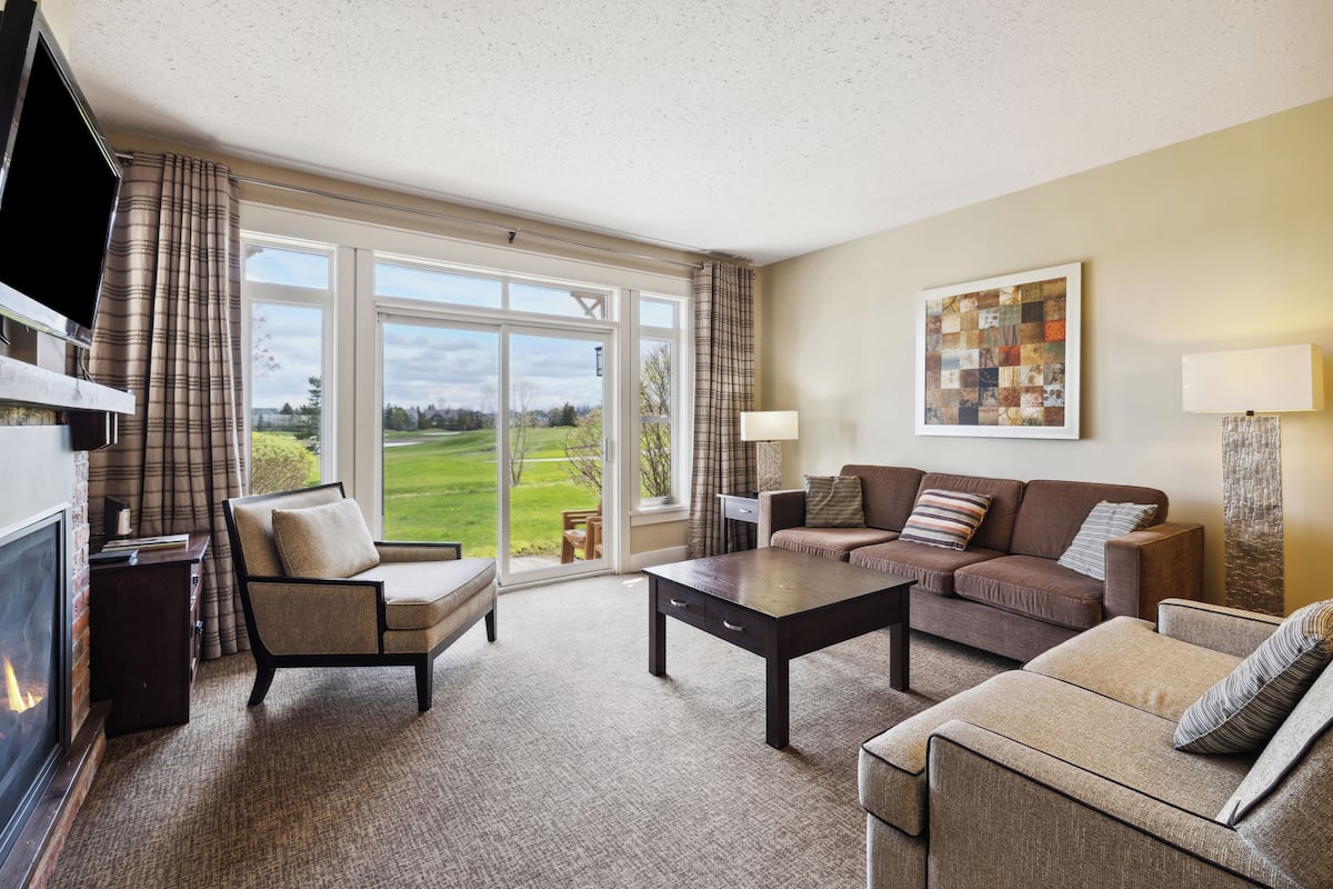 A spacious living room with a fireplace, a large window offering a view of a green landscape, a modern sofa set, and a flat-screen TV.