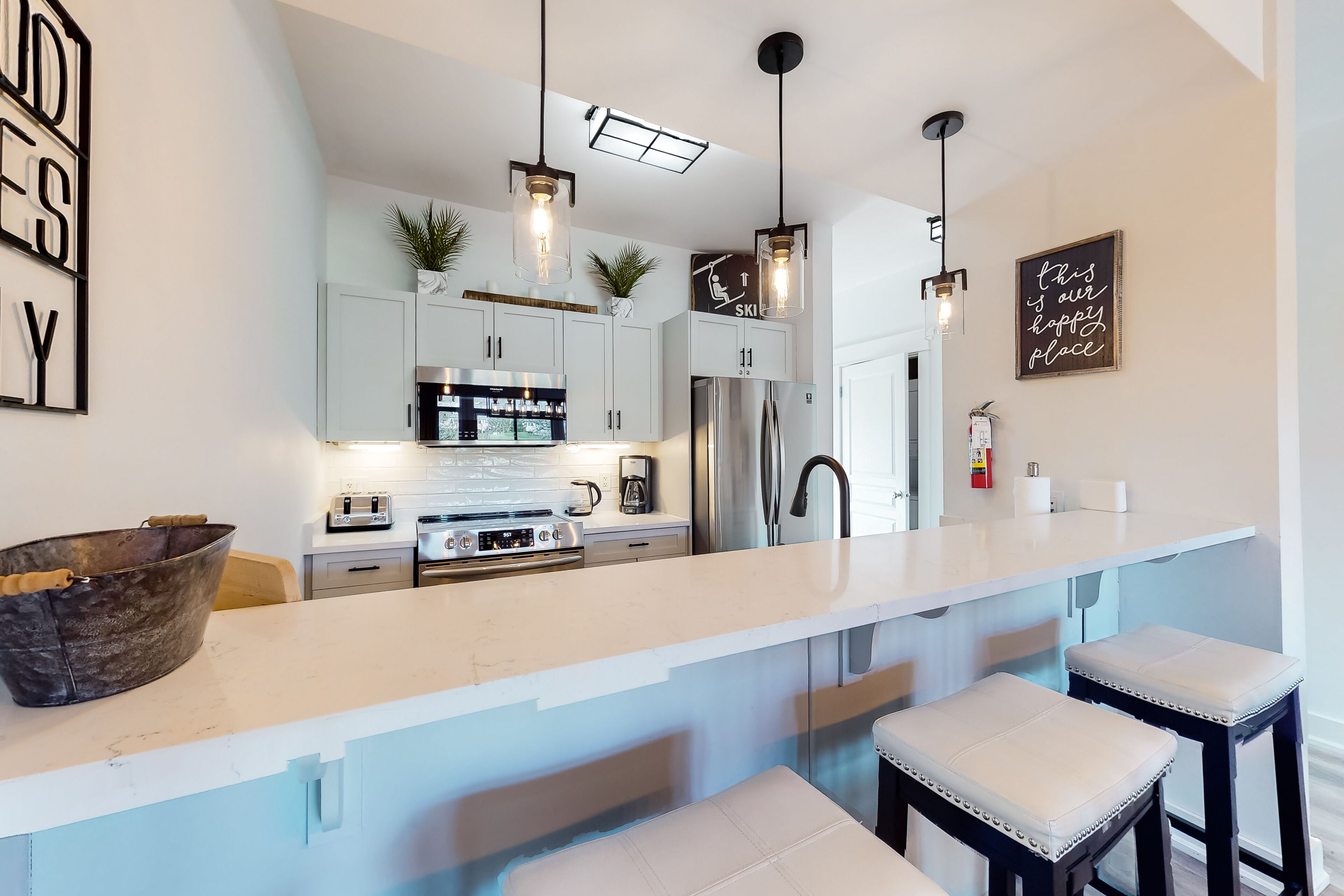 A modern kitchen with white cabinets, a large island with stools, pendant lighting, and a stainless steel refrigerator.