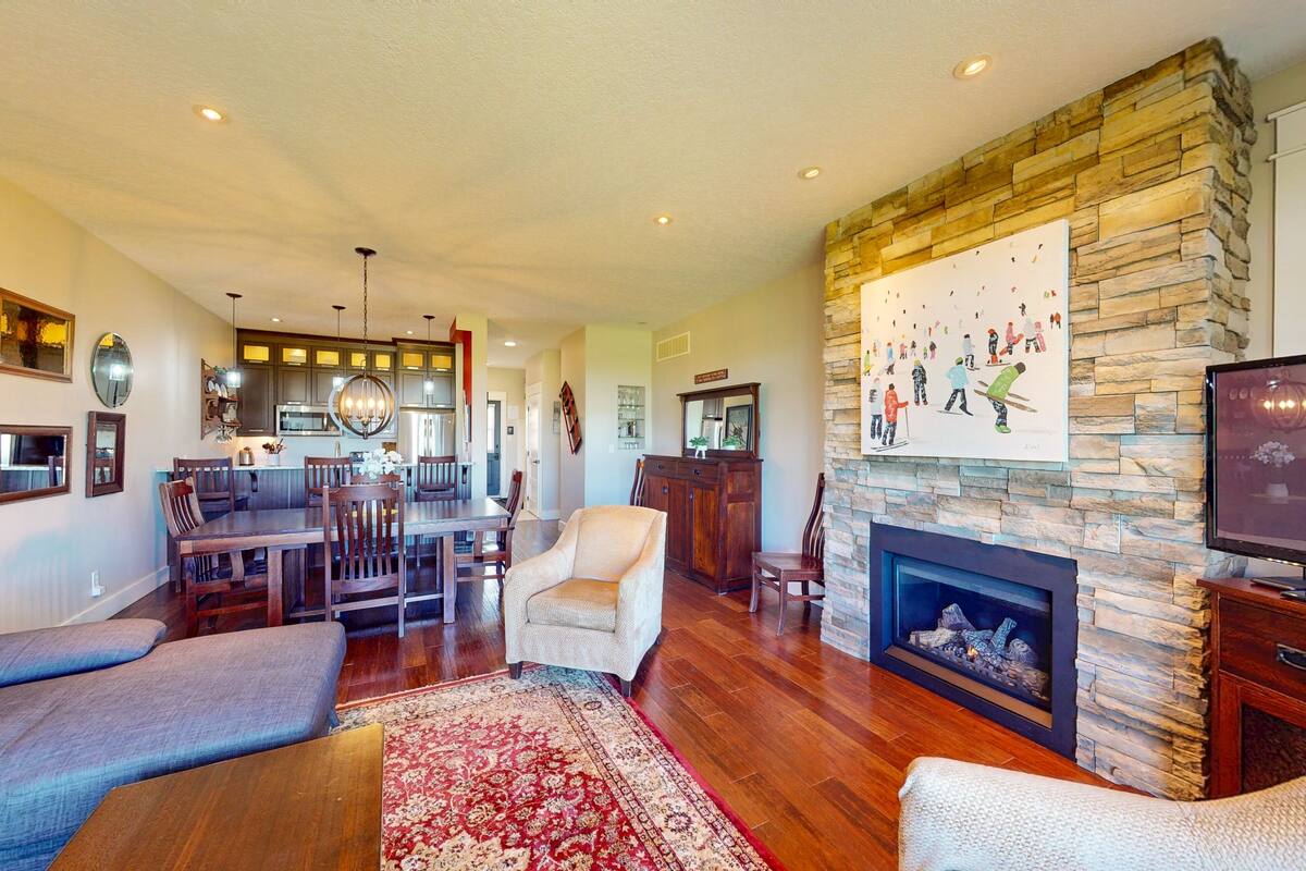 A spacious living room with a fireplace, a dining area, and a kitchen in the background.