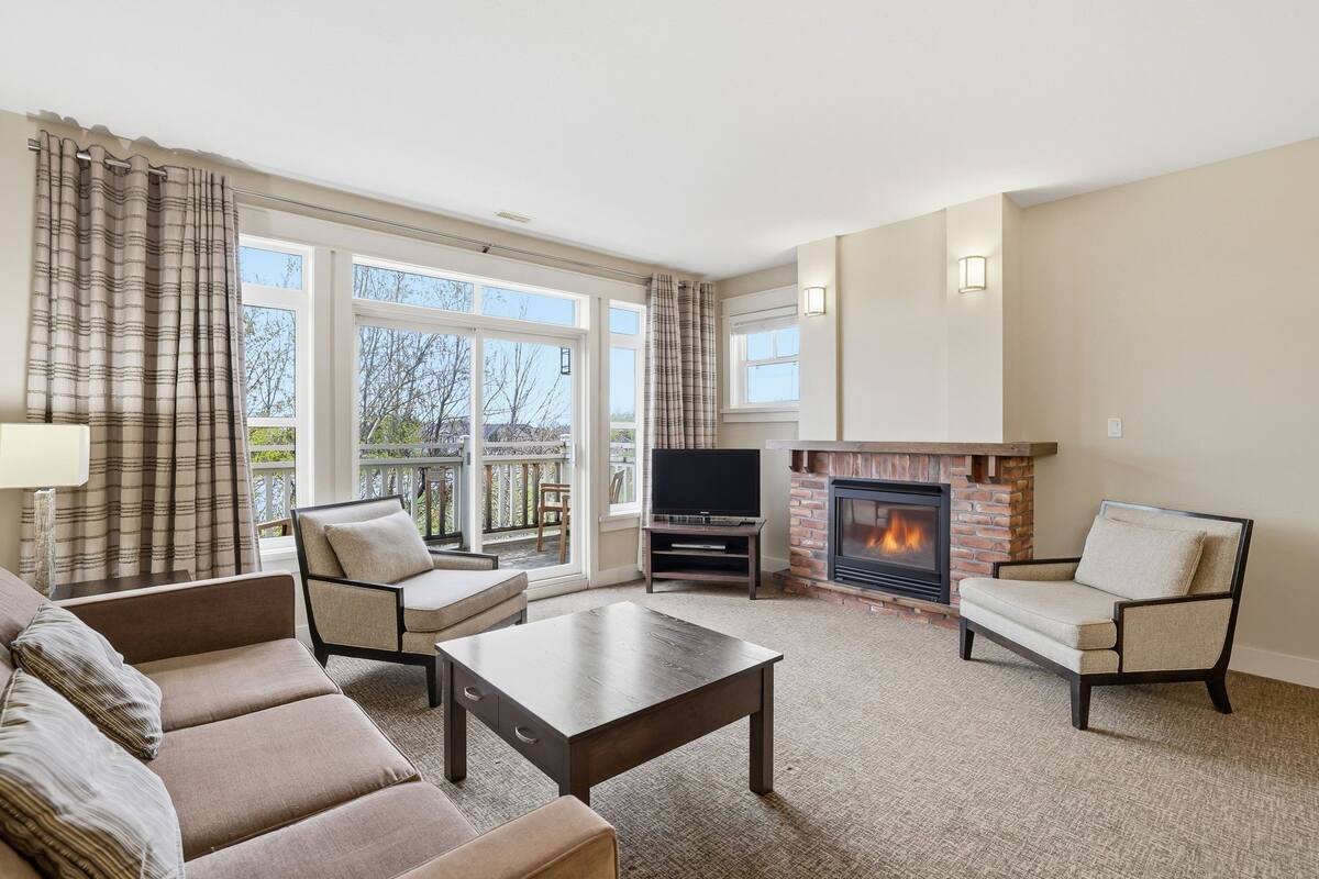 A living room with a sofa, chairs, a TV, a fireplace, and large windows.