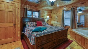 A wooden cabin bedroom with a bed, a nightstand, a lamp, a ceiling fan, and a large window. - Couples Getaway! Private, Romantic, Cabin with Jacuzzi & Hot Tub in Gatlinburg (Gatlinburg)