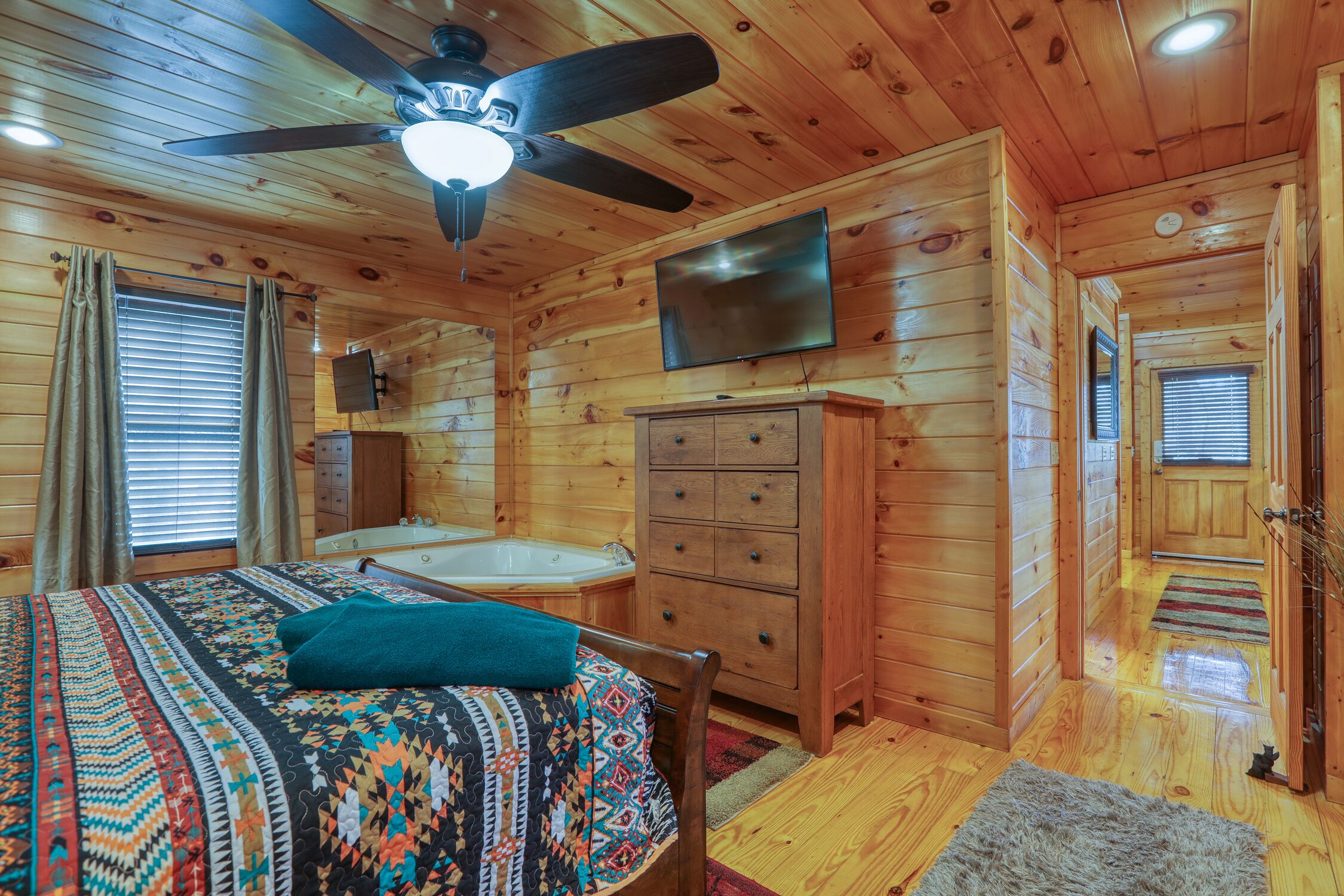 A wooden cabin room with a bed, a ceiling fan, a TV, a wooden dresser, and a bathtub.