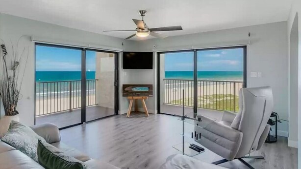 Uma sala de estar ampla com uma grande porta de vidro deslizante que dá para uma varanda com vista para a praia.