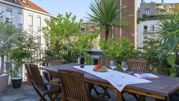 Une terrasse extérieure avec une table en bois dressée pour le repas, entourée de plantes en pot et d’un palmier.