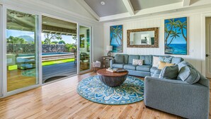 A spacious living room with a large sliding glass door leading to an outdoor area, a grey sofa set, a round coffee table, and tropical-themed artwork on the walls.