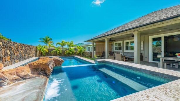 A swimming pool with a stone wall and palm trees, a covered patio with outdoor furniture, and a clear blue sky.
