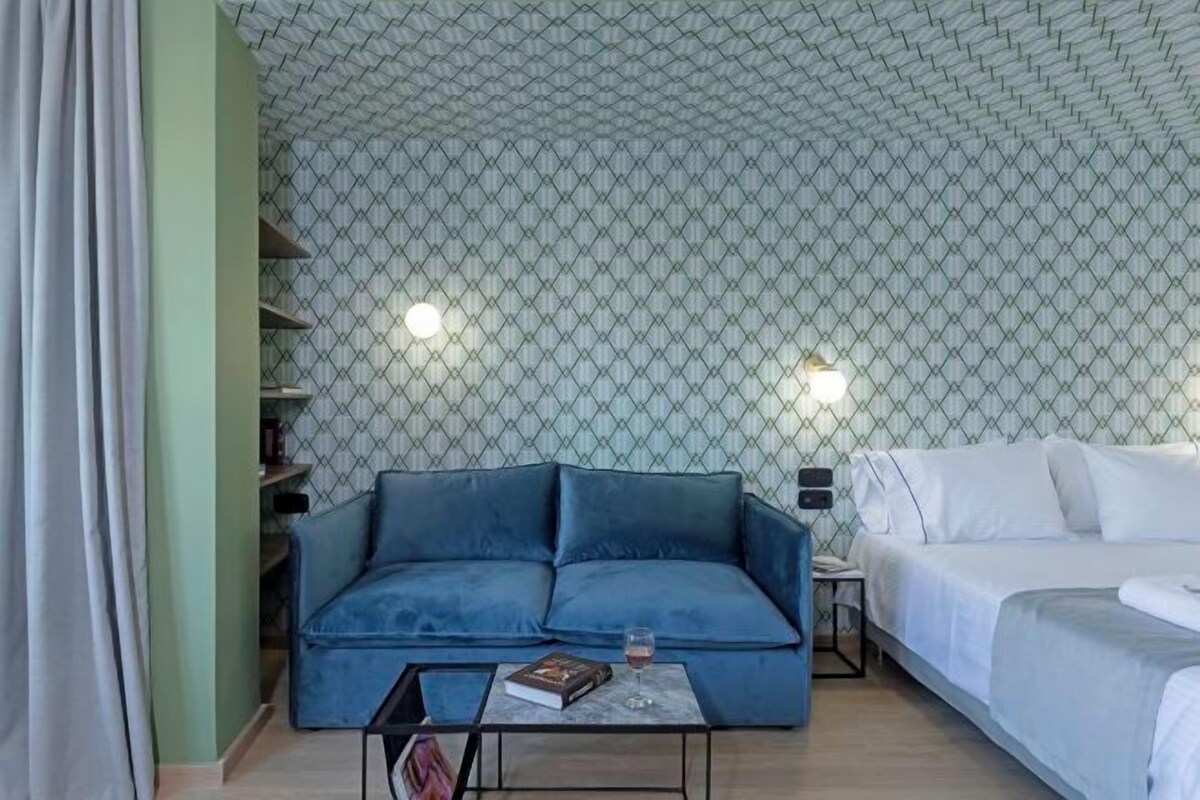 Una stanza con un divano blu, un letto, un tavolino con un libro e un bicchiere, e un soffitto decorato.