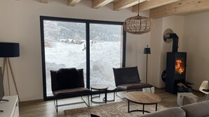Un saló d'estar acollidor amb una llar de foc, un sofà, una cadira i una vista d'un paisatge nevats a través de grans finestres.
