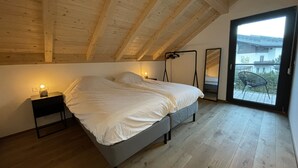 Un dormitori amb sostre inclinat, un llit, una tauleta de nit amb una làmpada i vista al balcó exterior.