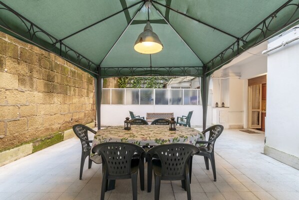 Une terrasse couverte avec une table et des chaises, un luminaire suspendu et une vue sur un immeuble avec des fenêtres.