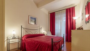 Ein Schlafzimmer mit einem Bett, einem Nachttisch mit Lampe, einer Kommode und einem Fenster mit roten Vorhängen.
