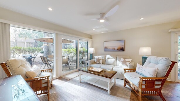 Un salon spacieux avec un canapé blanc, deux fauteuils, une table basse et une table à manger. De grandes fenêtres offrent une vue sur l’extérieur, et on peut apercevoir un ventilateur de plafond.