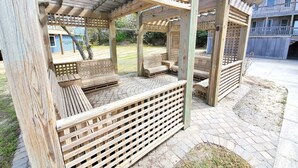 A wooden pergola with seating areas, a paved walkway, and a residential building in the background. - OBX Beach's, Natural Light, Roof Deck, Huge Home! (Kitty Hawk)