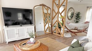 A living room with a TV on a white cabinet, a wooden chair, a hammock, and a large mirror on the wall. - OBX Beach's, Natural Light, Roof Deck, Huge Home! (Kitty Hawk)