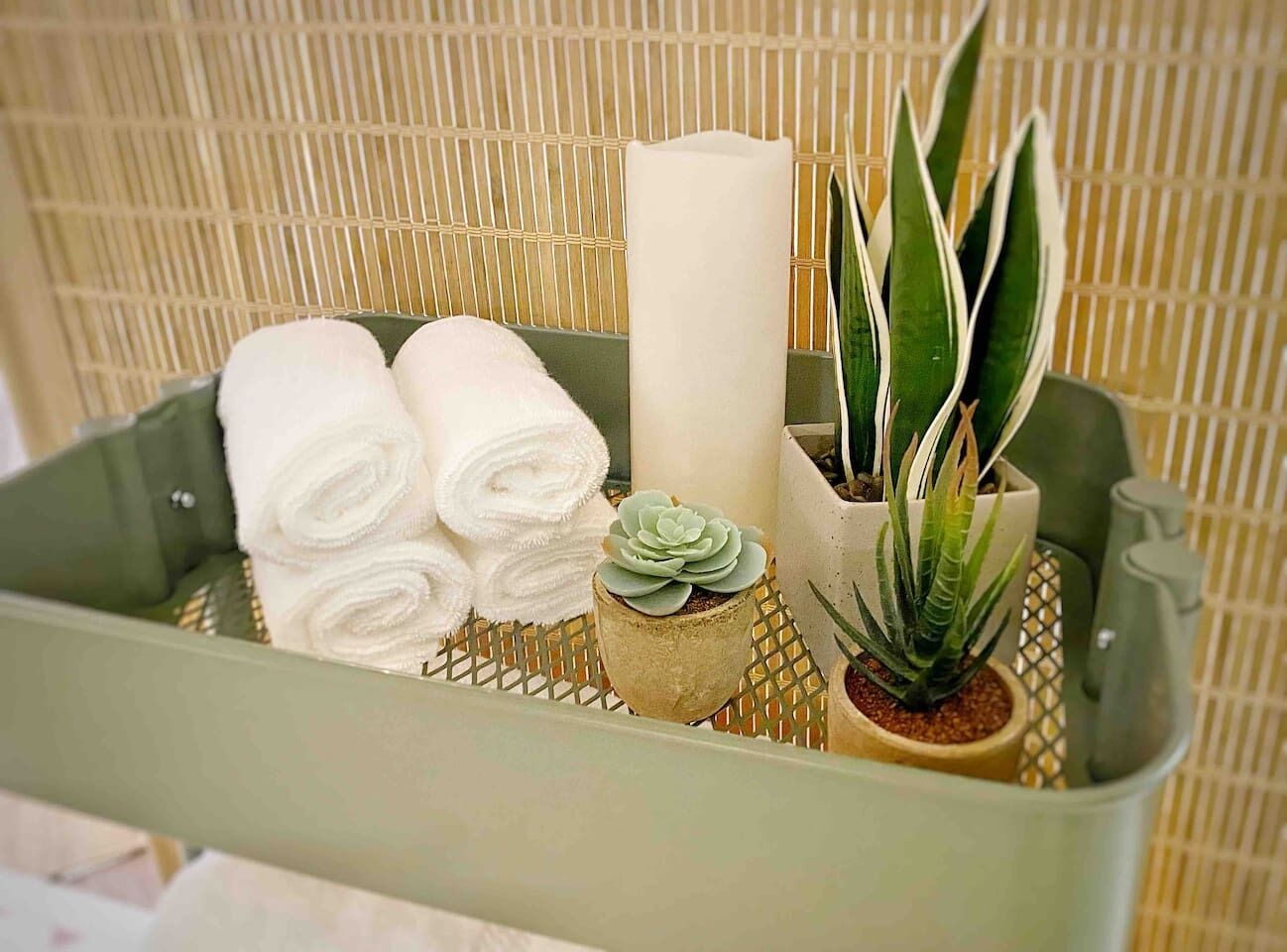 Una cesta con toallas blancas enrolladas, una vela blanca y dos plantas en macetas.