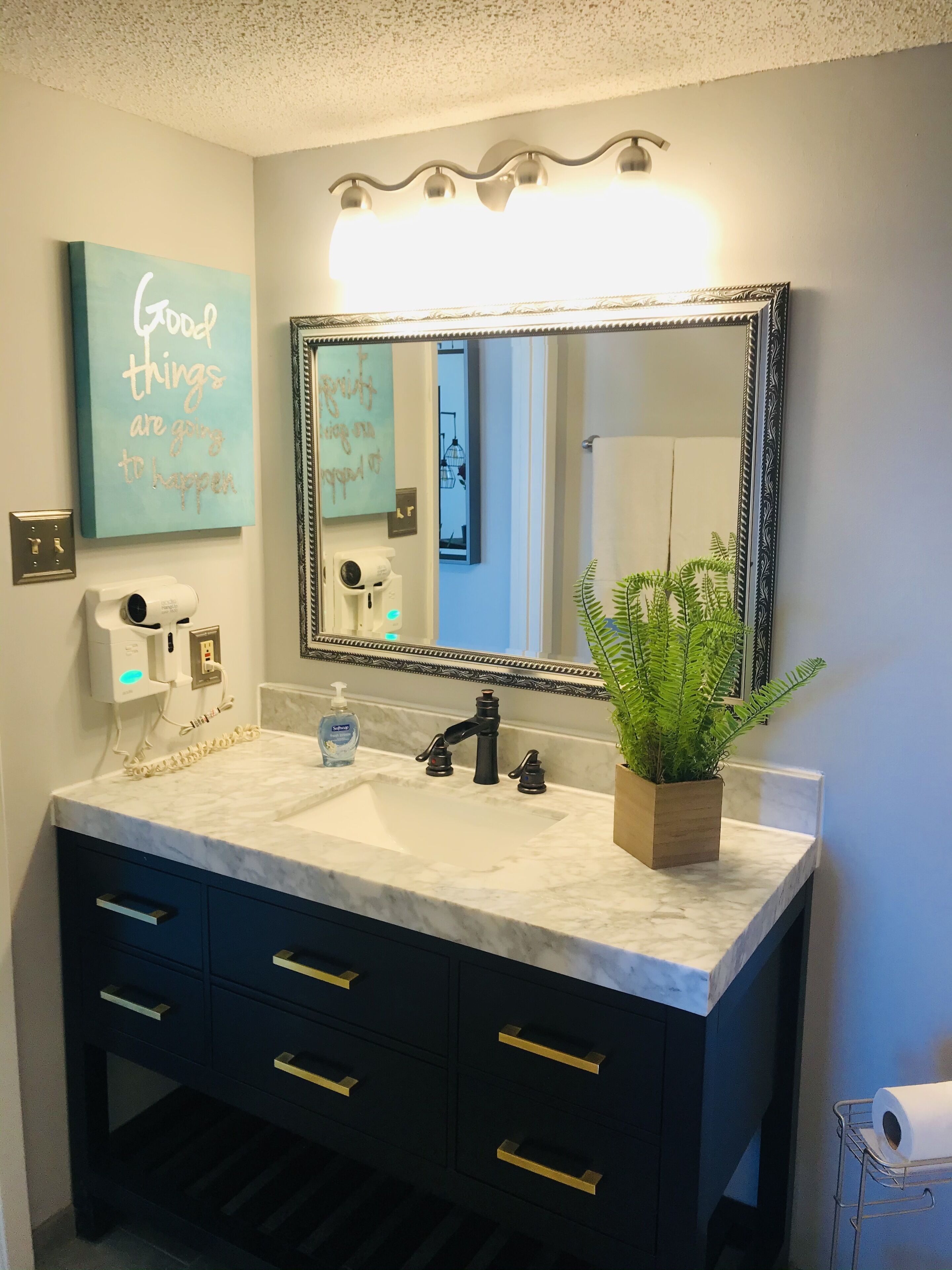 A bathroom with a marble sink, black cabinets, gold handles, a large mirror, a potted plant, and a motivational quote on the wall.