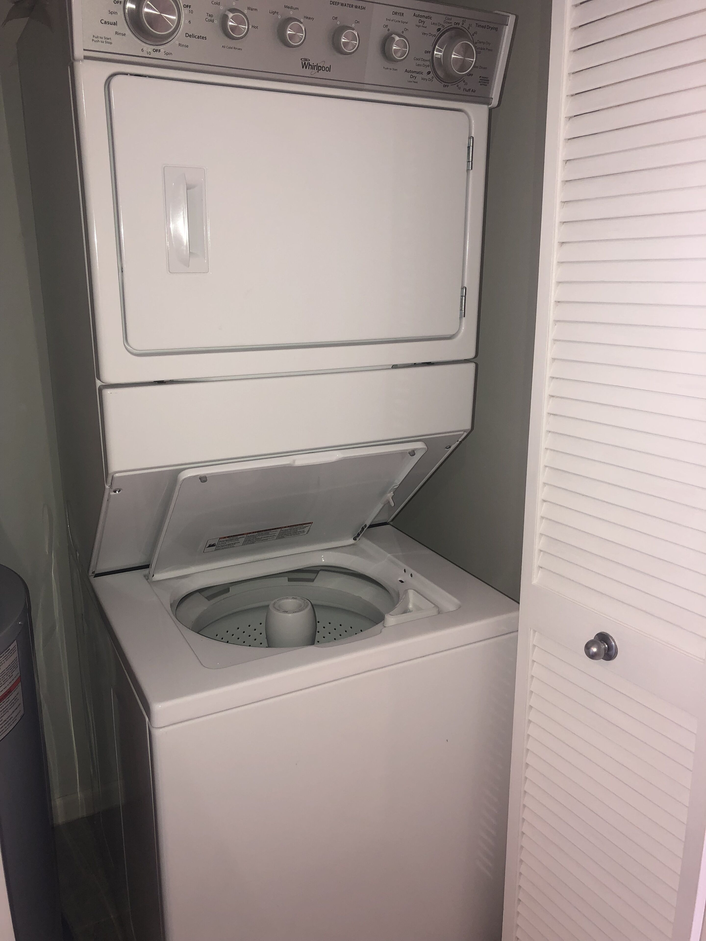 Una lavadora y secadora Whirlpool blanca y compacta, de carga frontal, ubicada en un lavadero pequeño con una puerta a la derecha.