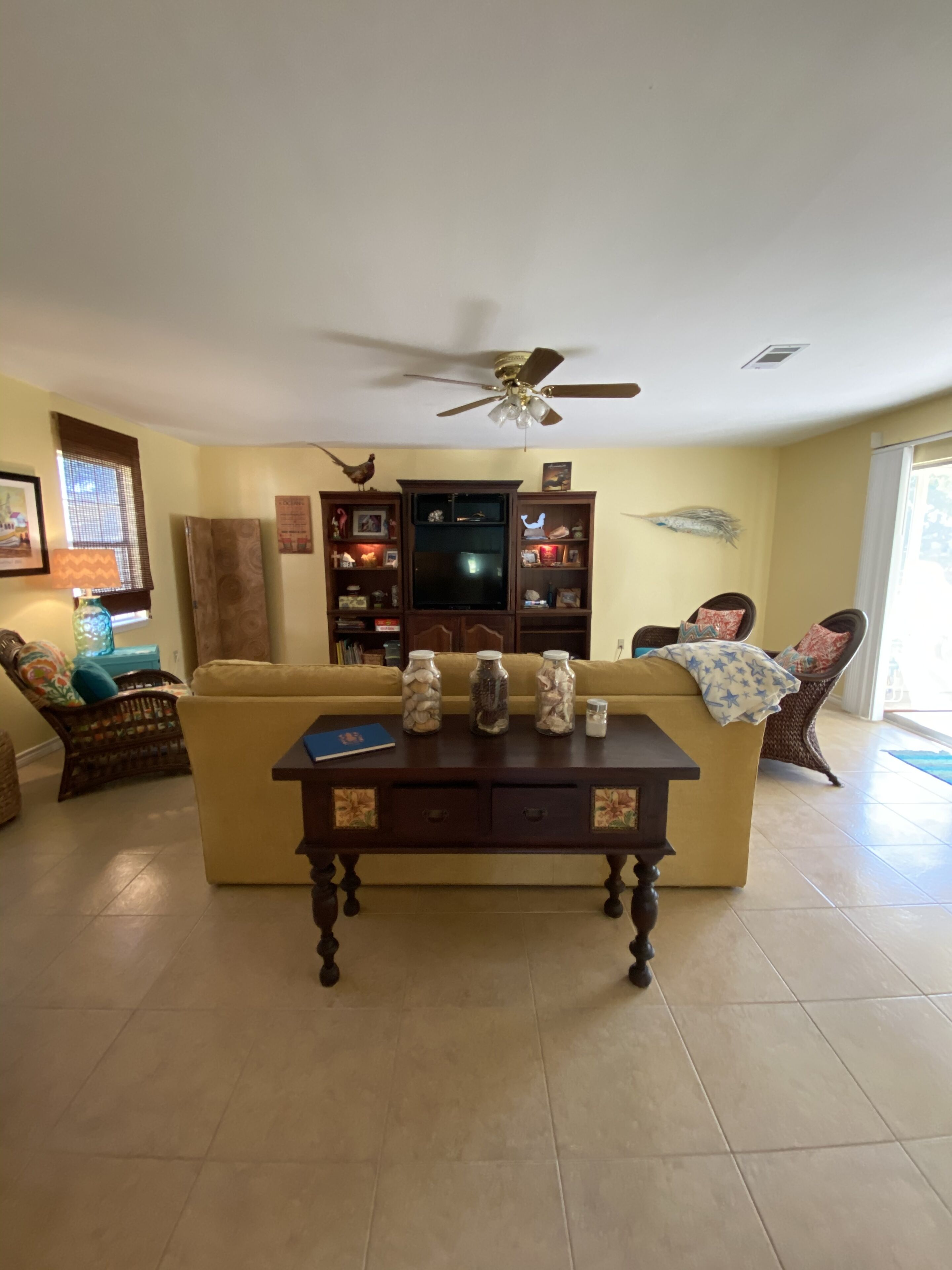 A living room with a yellow sofa, a wooden coffee table with decorative jars, a ceiling fan, and a TV stand with shelves.
