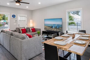 A modern living room with a grey sofa, a dining table set for four, and a large flat-screen TV displaying a beach scene.