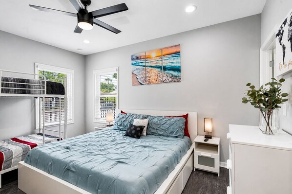 A bedroom with a bunk bed, a single bed, a ceiling fan, a painting of a beach, a bedside table with a lamp, and a vase with plants.