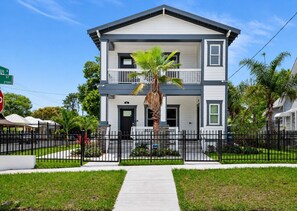 A two-story residential building with a front porch, a palm tree, and a black metal fence. - Ybor City Group Home, Sleeps 12 | Emperor Castle: Game Room & Parking (Tampa)