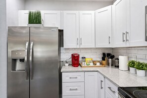 A modern kitchen with white cabinets, a stainless steel refrigerator, a coffee maker, and a countertop with various items.