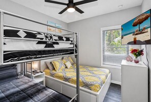 A bunk bed room with a ceiling fan, a TV on the wall, a window with blinds, and a bedside table with a lamp and decorative items.