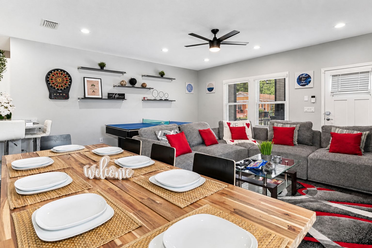 A modern living room with a ping pong table, grey sofas with red cushions, and a dining area with a wooden table set for six.