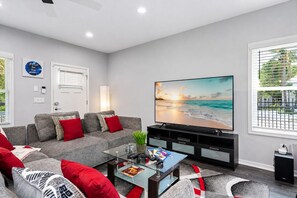A modern living room with a large flat-screen TV, grey sofa with red cushions, a glass coffee table, and a view of the beach through the window.