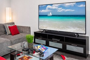 A living room with a large flat-screen TV displaying a beach scene, a grey sofa with red cushions, a glass coffee table with remote controls and a game controller, and a black entertainment unit.