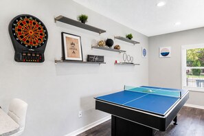 A room with a ping pong table, a dartboard, shelves with decorative items, and a window with a view. - Ybor City Group Home, Sleeps 12 | Emperor Castle: Game Room & Parking (Tampa)