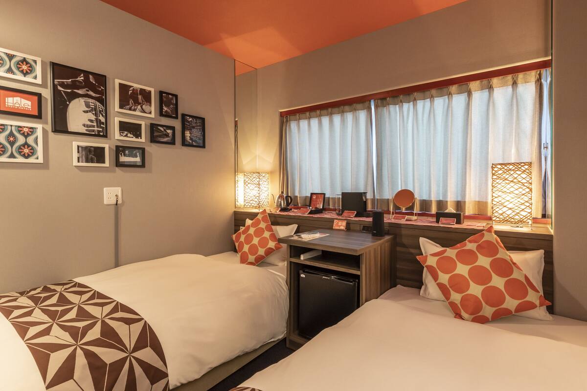 A hotel room with two beds, a desk, a refrigerator, and a window with curtains.