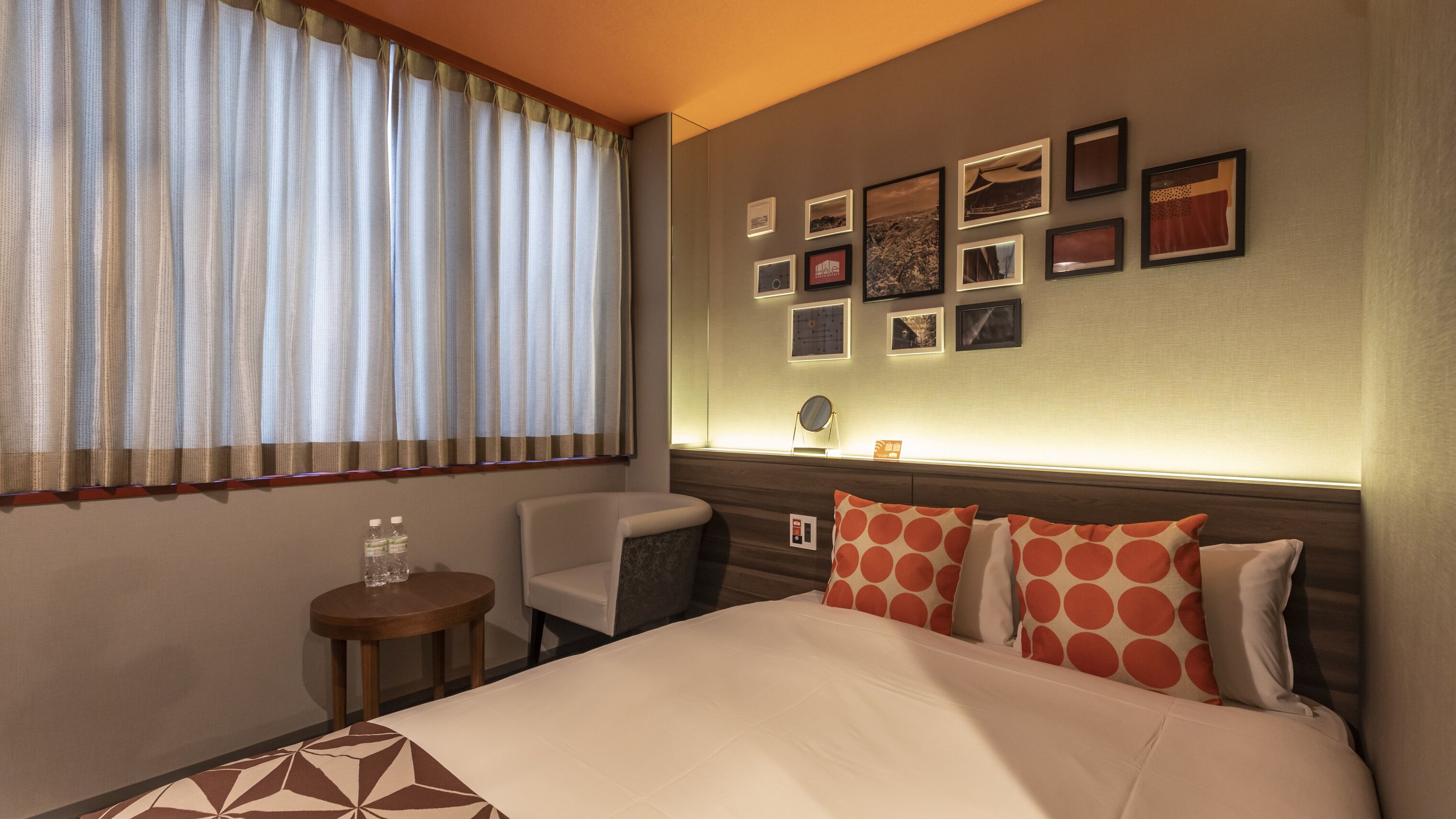 A hotel room with a bed, a chair, a small table, and a wall with framed pictures.