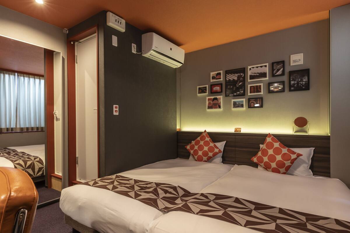 A hotel room with a bed, a chair, a wall-mounted air conditioner, and a series of framed pictures.
