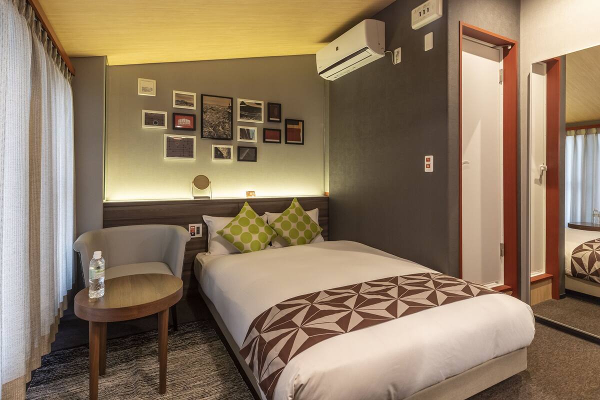 A hotel room with a bed, a chair, a small table, and a wall with framed pictures.