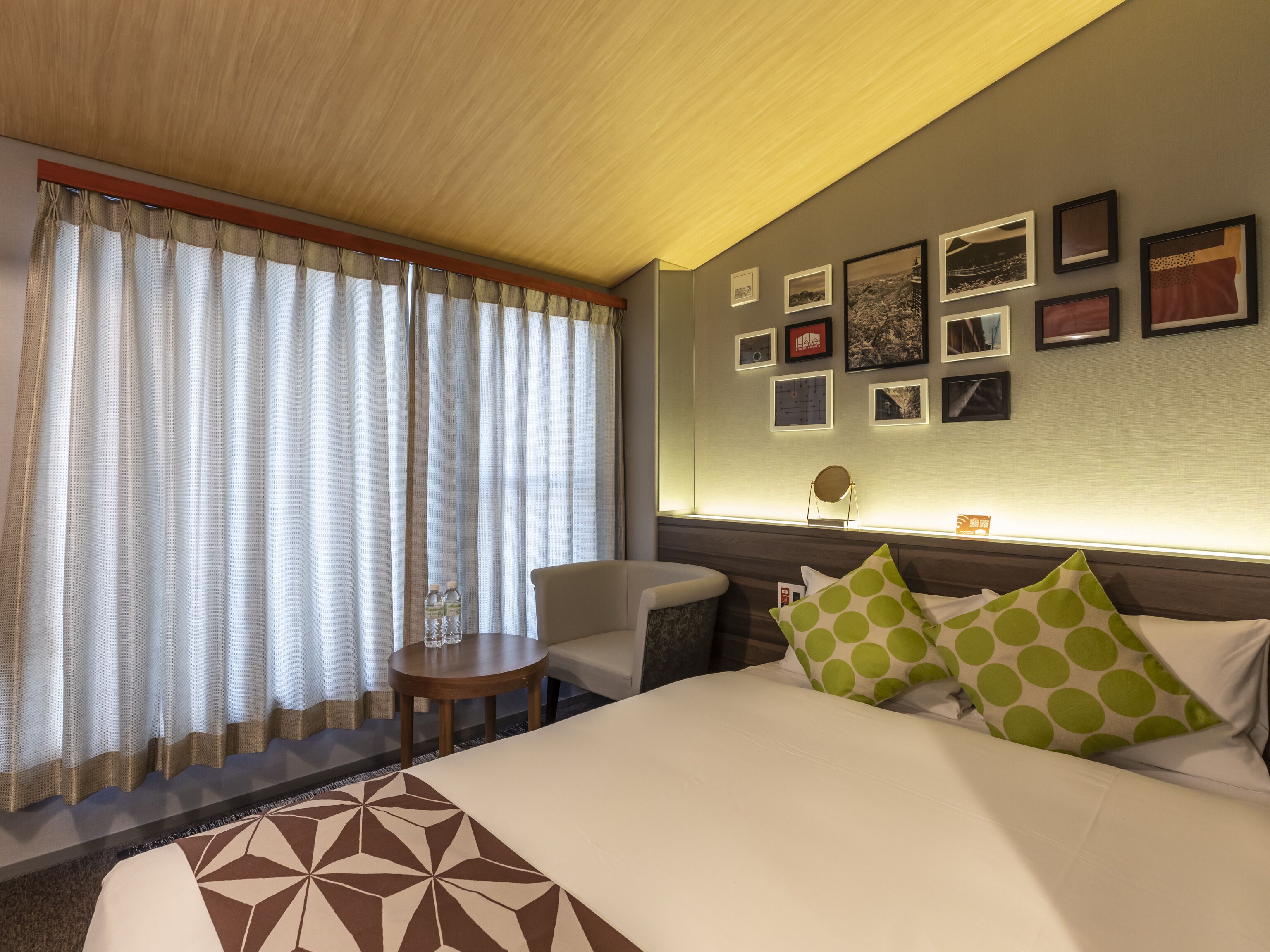 A hotel room with a bed, a chair, a small table, and a wall with framed pictures.
