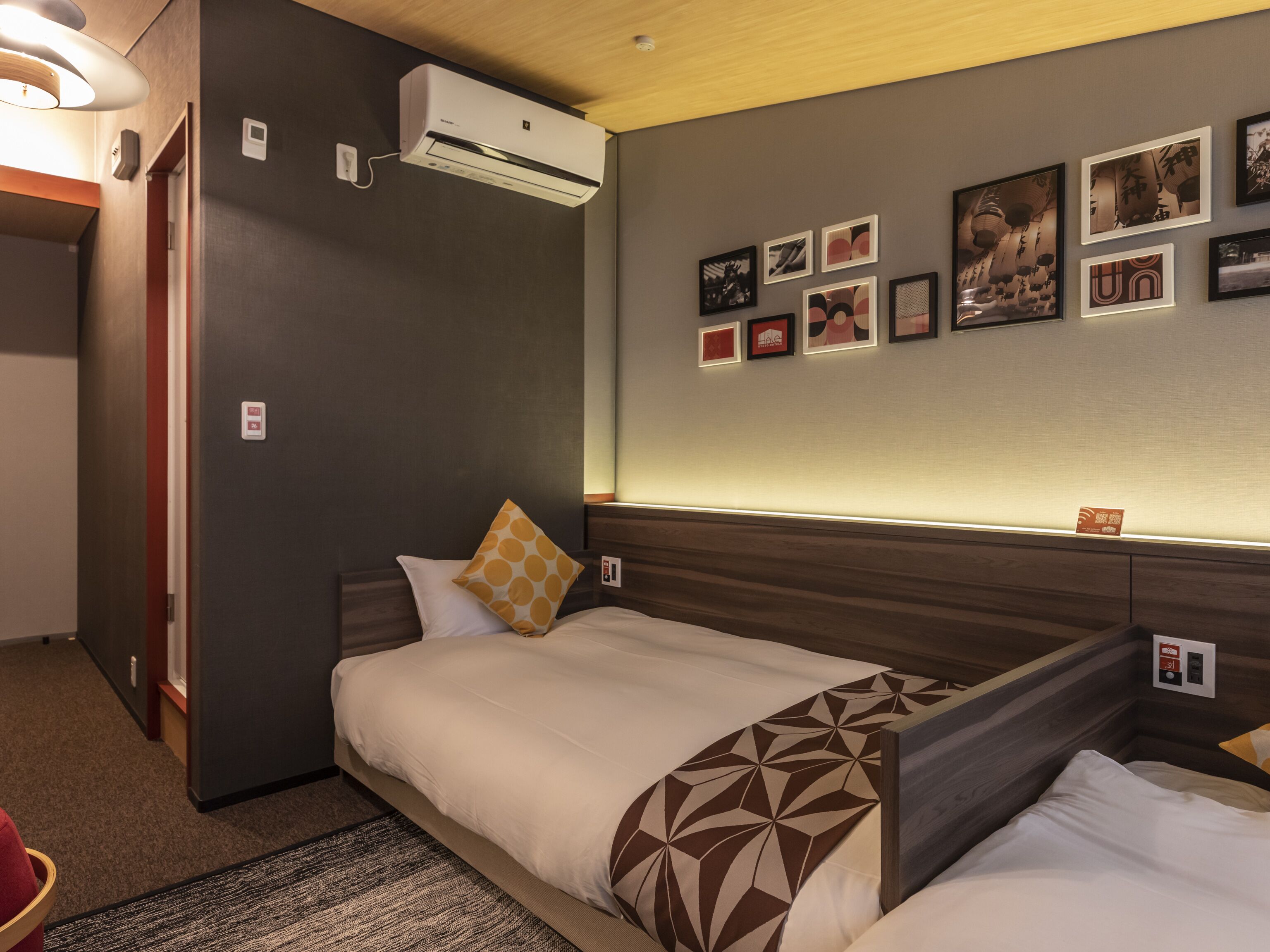 A hotel room with a bed, a wall-mounted air conditioner, and framed pictures on the wall.