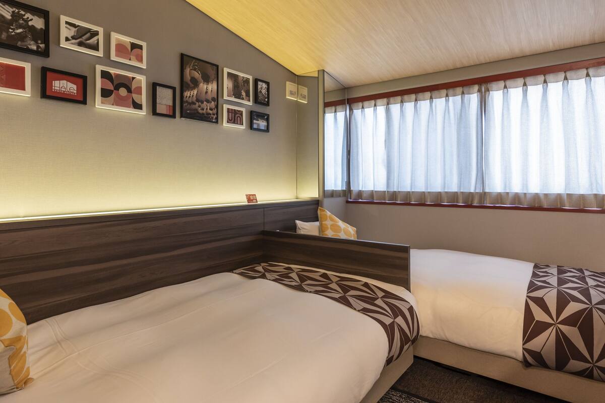 A hotel room with two beds, a large window with curtains, and a wall decorated with framed pictures.