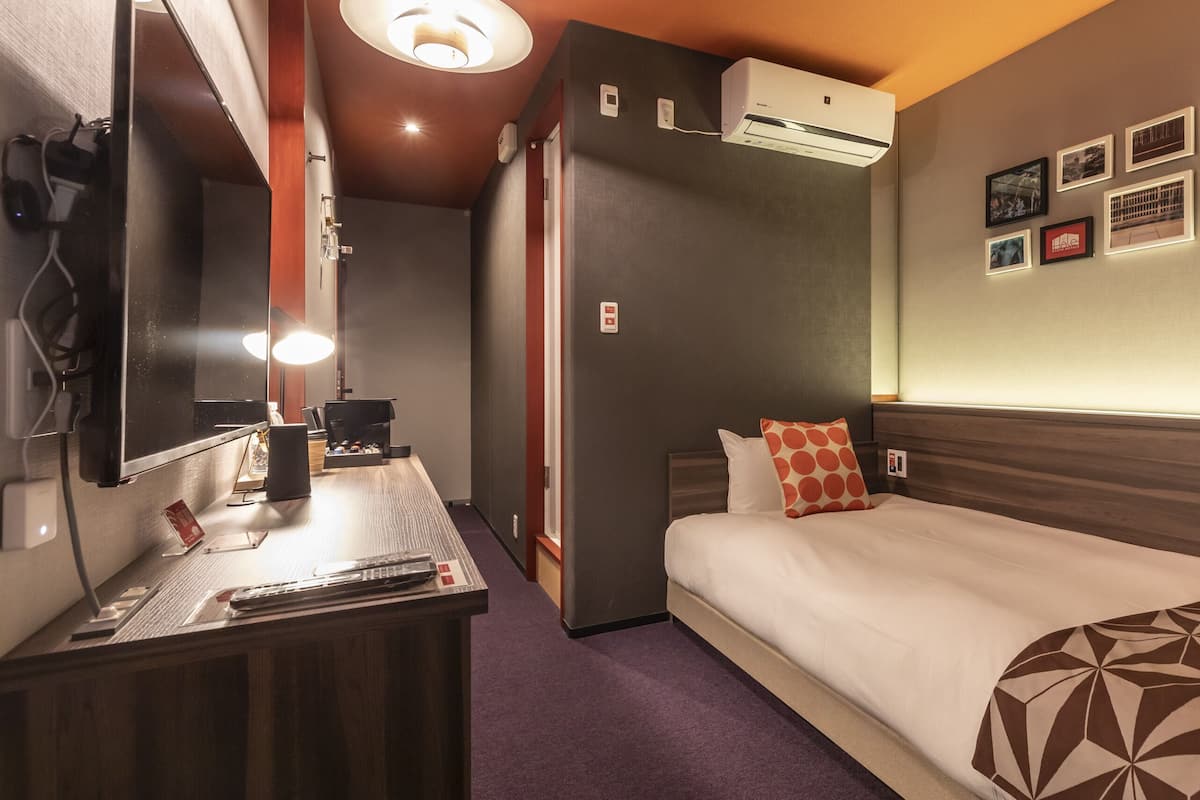 A hotel room with a bed, bedside table, lamp, air conditioning unit, and wall-mounted artwork.